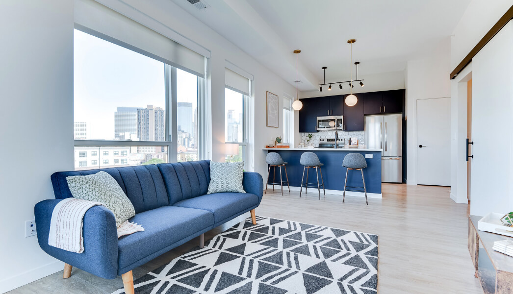 Modern living room with blue sofa, geometric rug, sleek kitchen, and panoramic city view through large windows.